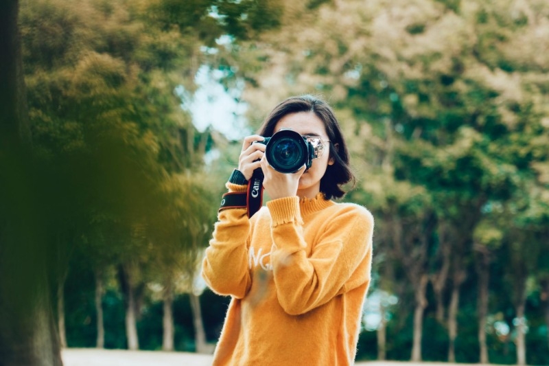 Woman in yellow sweater outdoors taking a photo with a camera.